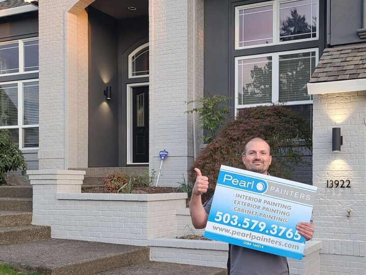 A painter stands in front of a Portland house holding a "Pearl Painters" sign advertising painting services.