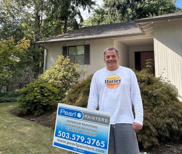 A man in a white Hurley shirt stands outside a house in Portland next to a Pearl Painters sign advertising painting services and phone number.