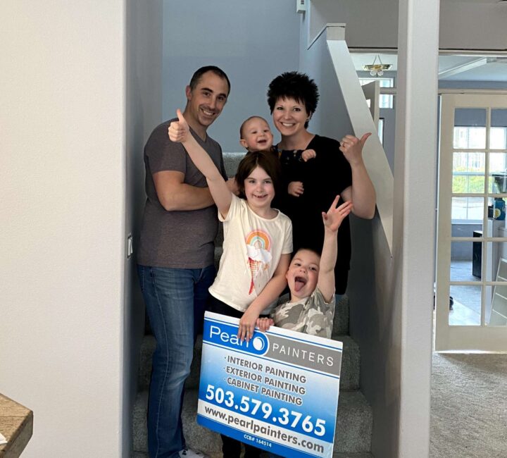 A family of five stands on indoor stairs in Portland, holding a Pearl Painters sign and giving thumbs up for painting services.