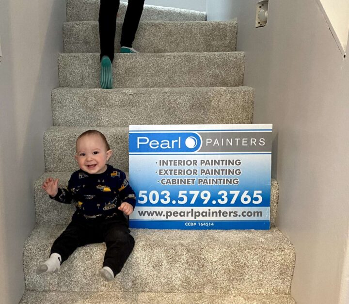 A baby sits on carpeted stairs in Portland next to a Pearl Painters sign with service and contact details, as someone walks down the stairs.