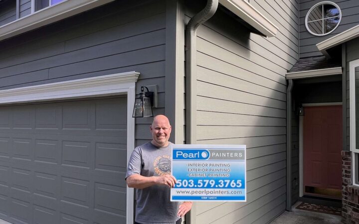 A painter stands in front of a gray house in Portland, holding a Pearl Painters sign offering interior, exterior, and cabinet painting services.