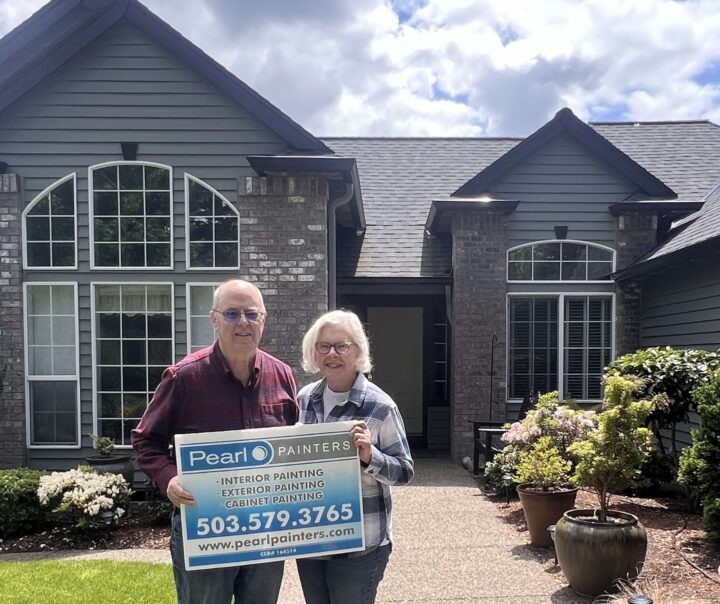Two people stand in front of a gray house in Portland holding a Pearl Painters sign advertising painter services for interior, exterior, and cabinets.