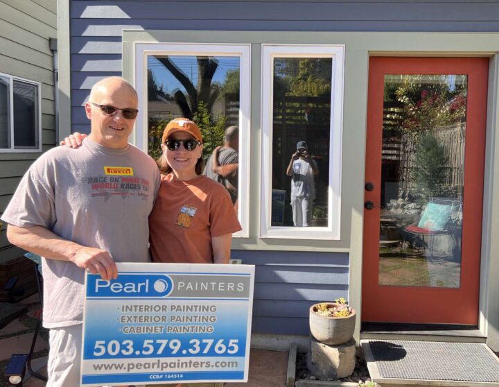 Two people stand outside a house in Portland holding a Pearl Painters sign advertising their painting services. The photographer is visible in the window reflection.