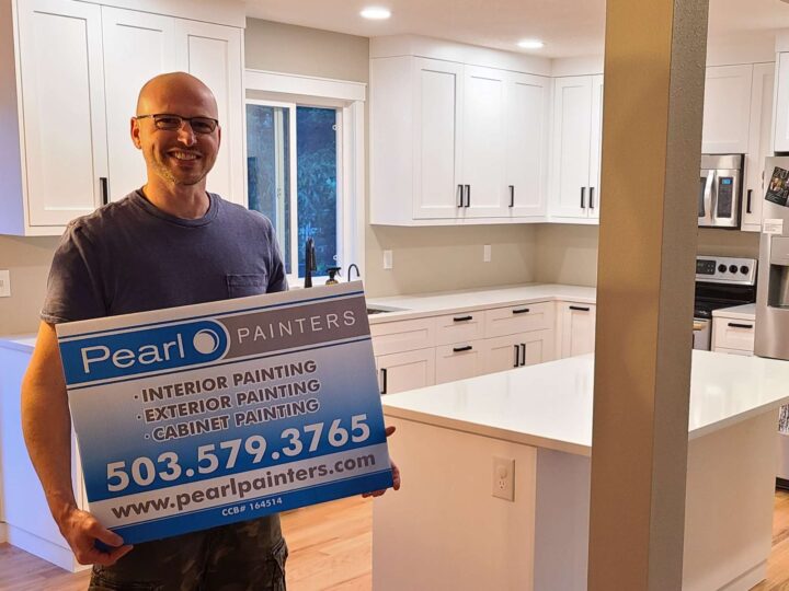 A painter stands in a modern white kitchen in Portland holding a Pearl Painters sign promoting interior, exterior, and cabinet painting services with contact details.