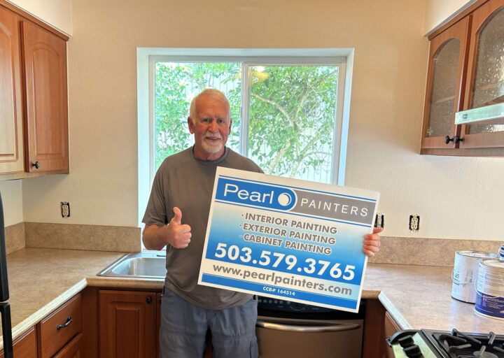 An older man in a kitchen holds a Pearl Painters sign with the company’s painting services and contact details. He gives a thumbs up, representing the Portland painter company.