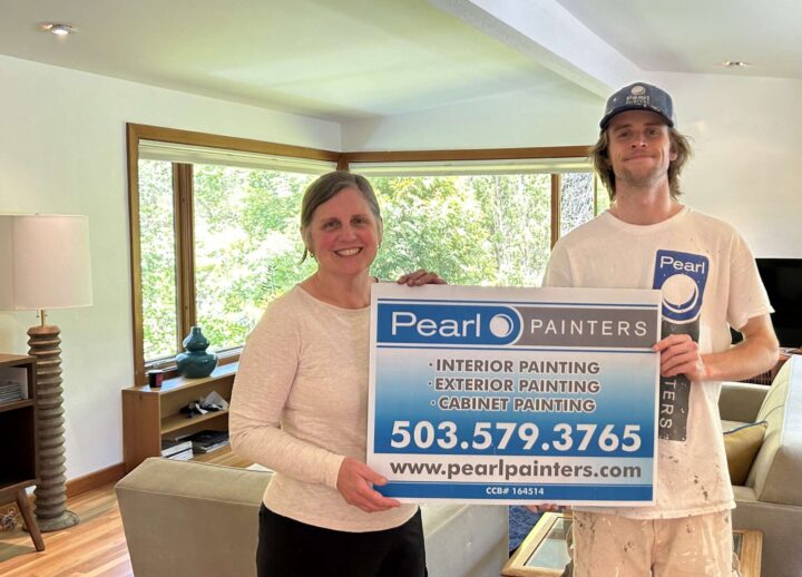 Two people stand indoors in a modern room with large windows, holding a Pearl Painters sign advertising interior, exterior, and cabinet painting in Portland.