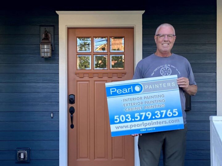 A painter stands in front of a blue house in Portland holding a Pearl Painters sign with services and phone number.