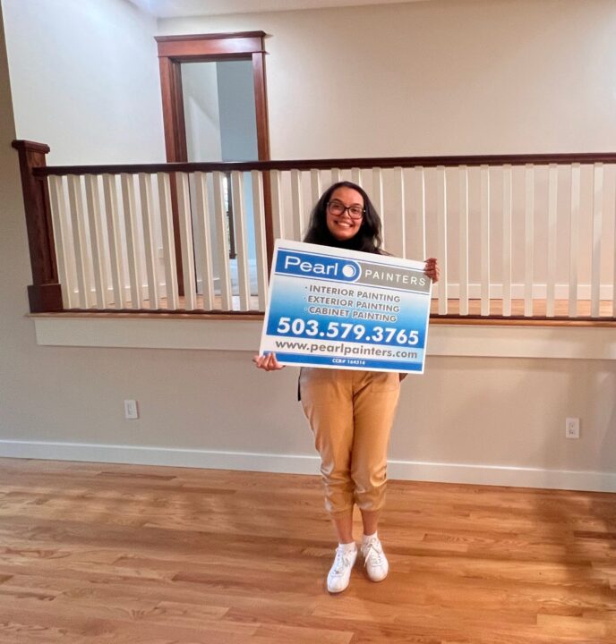 A painter stands indoors in Portland holding a Pearl Painters sign that lists contact info and interior, exterior, and cabinet painting services.