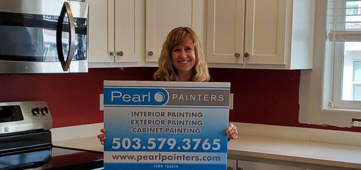 A woman in a kitchen holds a Pearl Painters sign advertising painter services—interior, exterior, and cabinet painting—in Portland.