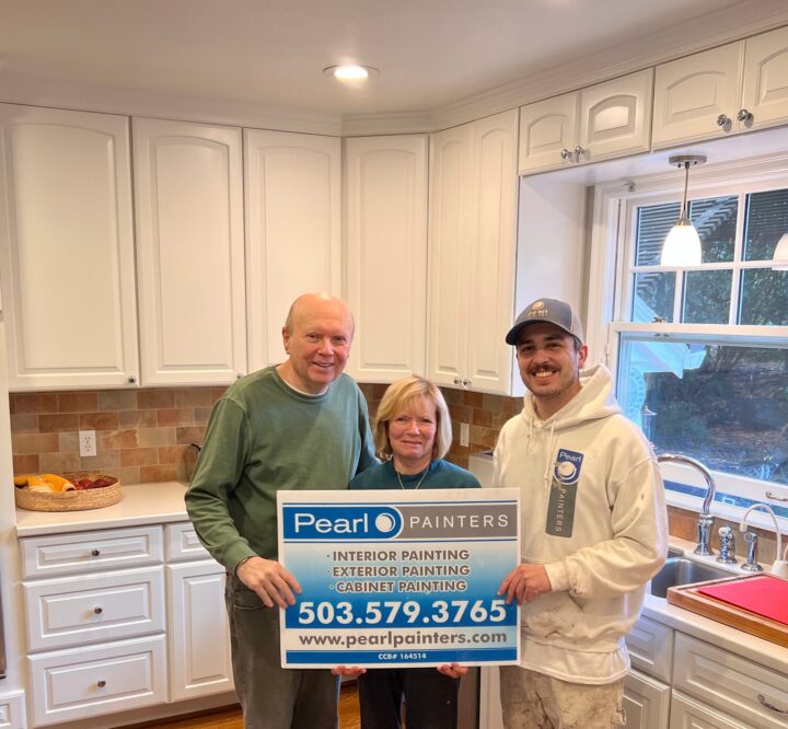 Three people stand in a kitchen in Portland holding a Pearl Painters sign advertising their painting services. White cabinets and a window are visible.