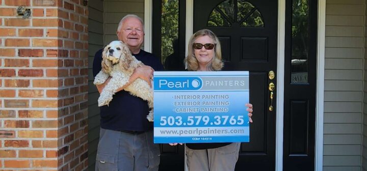 Two people stand by a house door in Portland, one with a dog. They hold a "Pearl Painters" sign advertising local painting services.