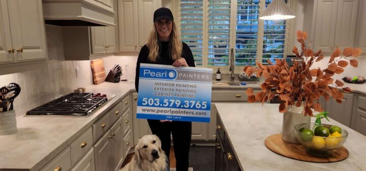 A woman stands in a Portland kitchen holding a Pearl Painters sign. A dog sits at her feet. The painting scene shows white cabinets, a marble countertop, and an island with a vase of orange leaves.