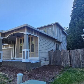 A two-story beige house with white trim, arched front porch, and a wooden fence on the right side, set against a clear blue sky.