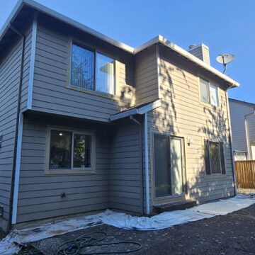 A two-story house with gray siding and several windows. The backyard has a partially covered ground with a hose and plastic sheets, and is fenced-in with neighboring homes visible in the background.