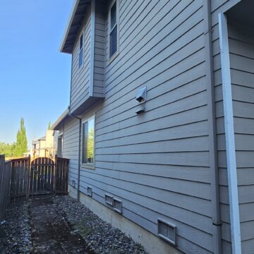 Side view of a house with grey siding, a narrow garden path, and a wooden fence. The soil near the path appears to have been recently worked on. Clear blue sky in the background.