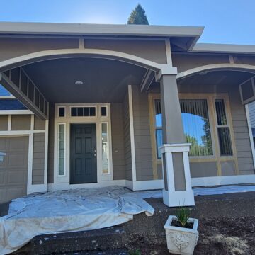 A two-story house with a large covered front porch featuring two arched entrances. The house is painted in beige tones with white accents and has double garage doors on the left.