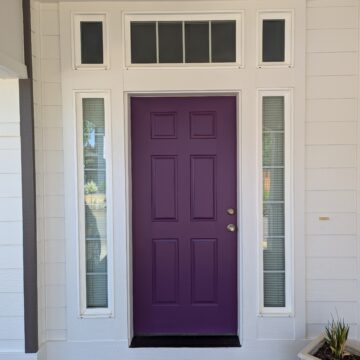 A white house entrance with a purple front door. The door is flanked by two tall, narrow windows with blinds. A potted plant is on the right side of the entrance.