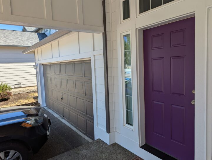 A house entrance with a purple front door and an adjacent garage door. A dark-colored car is partially visible in the driveway. White and gray exterior walls with a narrow window next to the entrance.