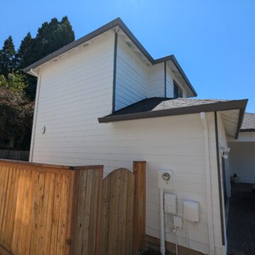 A two-story white house with a wooden fence on the side and a tree in the background. The house has a slanted roof and visible utility connections on the outside wall.