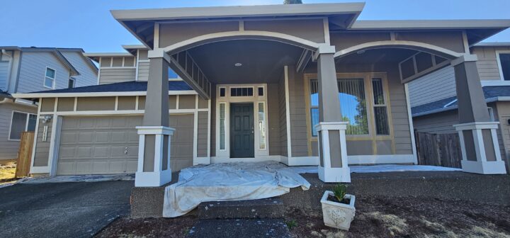 Front view of a two-story house with a covered porch, a dark green front door, and a garage. Plastic covers part of the porch area, indicating possible ongoing painting or remodeling work.