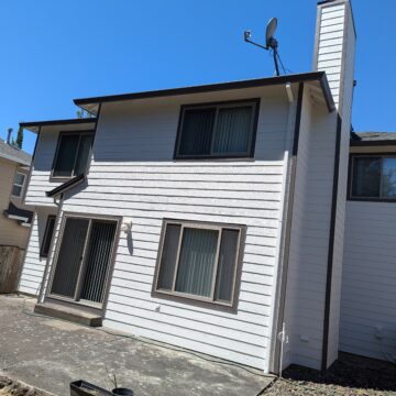 A two-story house with white siding, brown trim, and a satellite dish on the roof. The patio area is visible in the foreground. The sky is clear and blue.