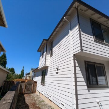 A white two-story house with dark trim, adjacent to another similar structure, viewed from a narrow side yard on a clear, sunny day. A wooden fence separates the properties.