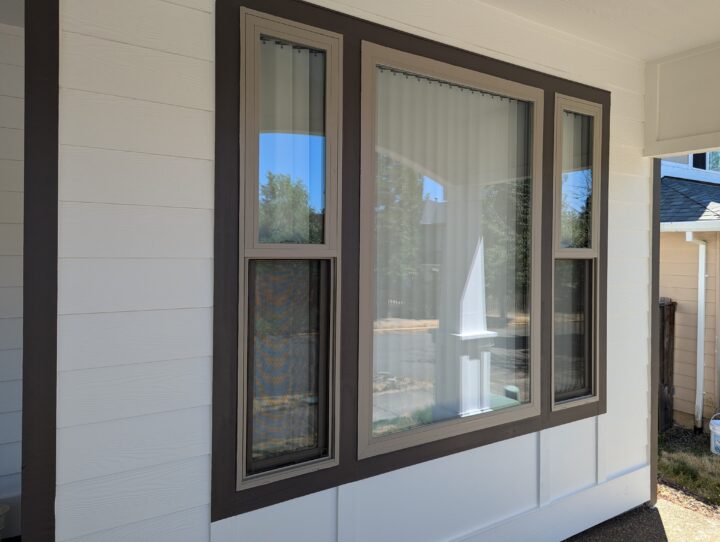 A large window with dark brown trim is set into the white exterior wall of a house. The window consists of one large central pane and two smaller side panes, all with vertical blinds.