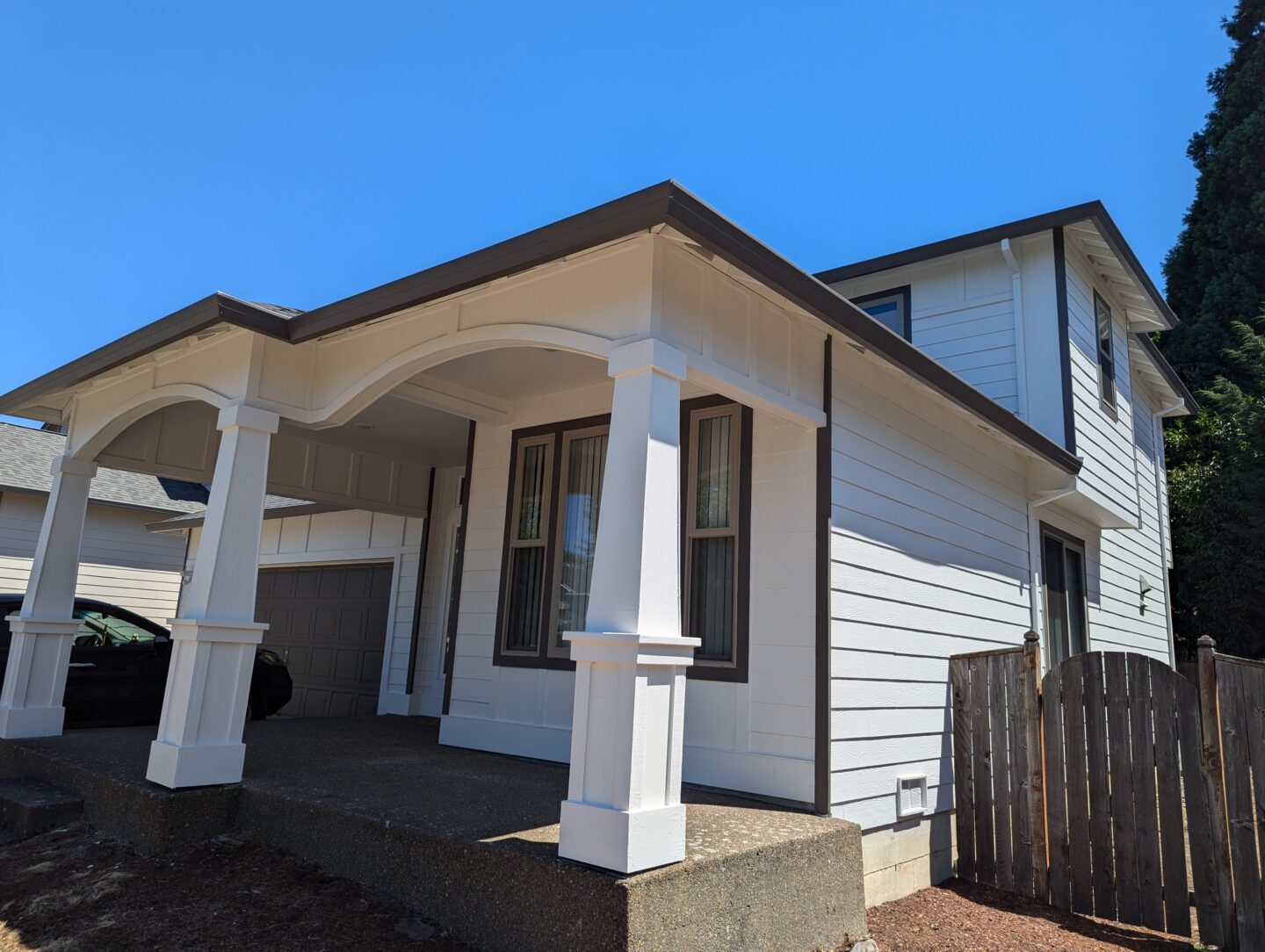 A two-story house with white siding, brown trim, and an attached carport. The front features an arched porch and a side gate leading to a fenced backyard.