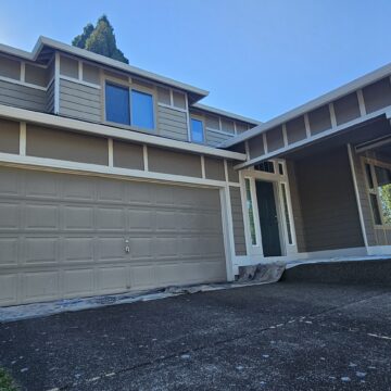 Two-story house with gray siding, white trim, and a two-car garage. The front yard features a concrete driveway with a slight slope.