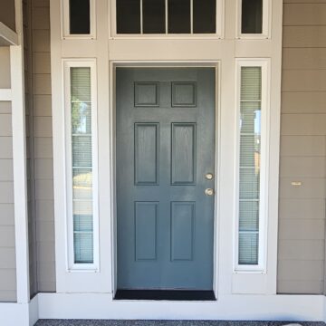 A gray front door with four panels and a chrome handle, flanked by narrow windows on both sides and a transom window above. The entryway is covered with a drop cloth.