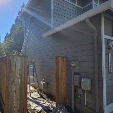 A side view of a house with light gray siding and a sloped roof. A ladder leans against the house, and a white tarp covers the ground nearby. A wooden fence is visible in the foreground.