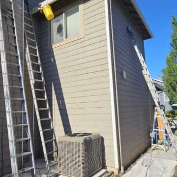 A small gray house extension with ladders leaning against it, paint supplies on the ground, and an air conditioning unit next to the building.