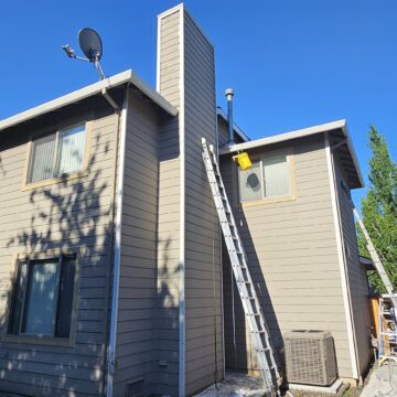 A two-story house with beige siding and a chimney, featuring a satellite dish. A ladder is propped up against the side of the house. Adjacent houses and clear blue sky are visible.