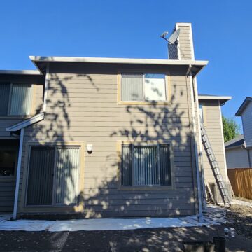 A two-story beige house with a sloping roof. There's a ladder leaning against the right side of the house, and a white tarp spread on the ground below. Fence and trees are visible in the background.