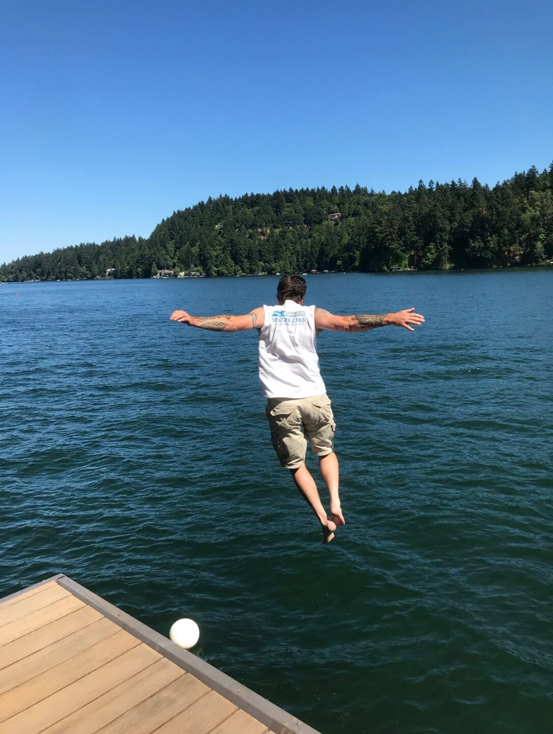 A person in a white tank top and shorts jumps off a dock into a lake, with their arms outstretched. Forested hills and a clear blue sky are in the background.