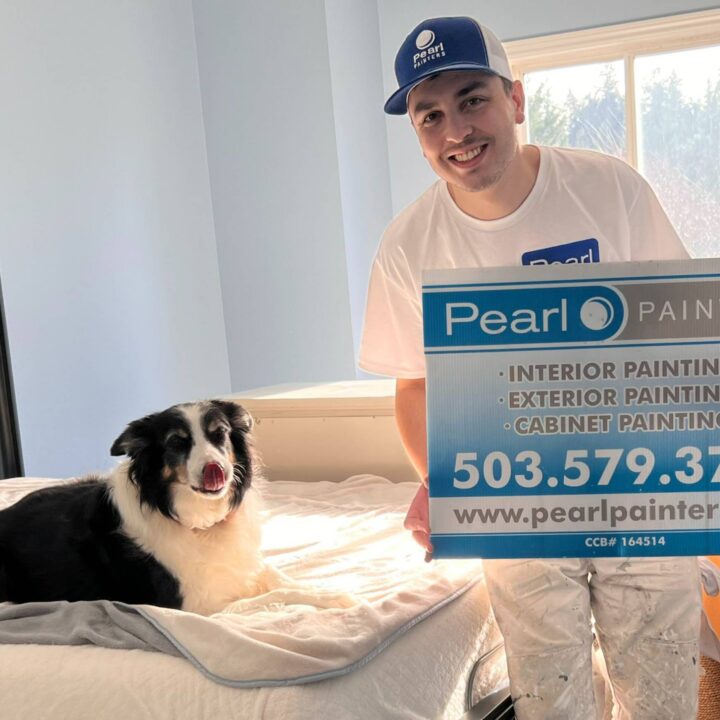 A painter in work clothes holds a Pearl Painters sign beside a black and white dog on a bed in a sunlit Portland room.
