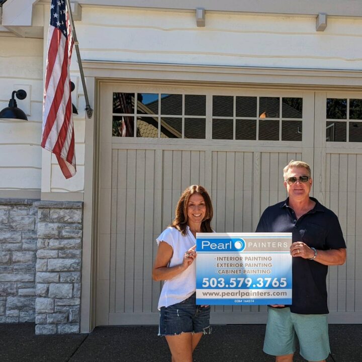 Two people hold a Pearl Painters painting sign in front of a garage. An American flag is on the nearby house. Likely Portland.
