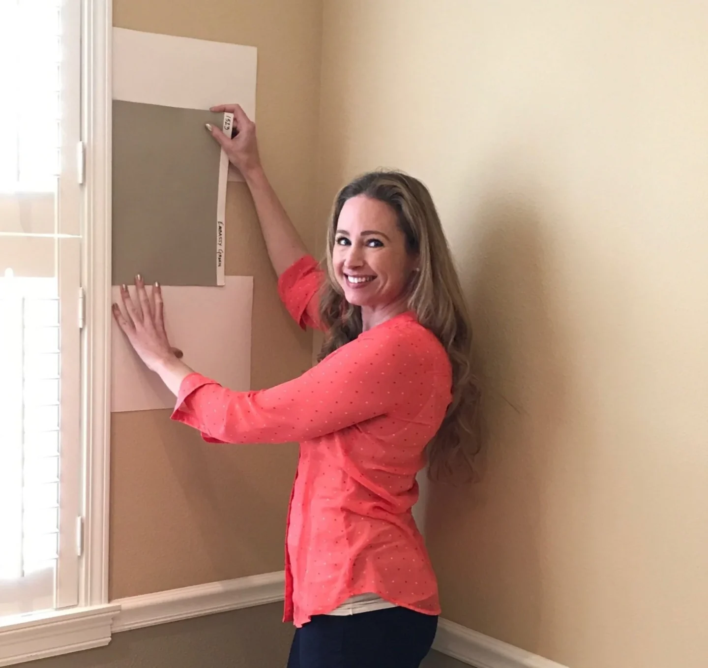 A painter in a coral blouse compares paint samples on a beige wall by a window, selecting colors for Pearl Painters in Portland.