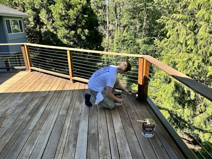 In June 2025, a painter stains the railing of the Shainsky home’s wooden deck in Portland. Nearby are a can of wood stain and a paint tray. Trees and the house are visible in the background, showing an outdoor painting project by Pearl Painters.