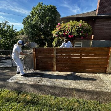 In May 2025, two painters in protective suits use a sprayer to apply stain or paint to an exterior wooden fence beside a house in Portland, with a tarp on the ground—possibly as part of a Pearl Painters or Trujillo painting project.