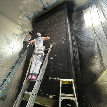 Two painters from the Turk team paint a tall black wooden interior structure in a room covered with plastic sheets. The renovation, managed by Pearl Painters in Portland, is scheduled for completion in May 2025.