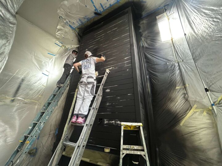 Two painters from the Turk team paint a tall black wooden interior structure in a room covered with plastic sheets. The renovation, managed by Pearl Painters in Portland, is scheduled for completion in May 2025.