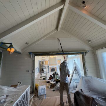 In May 2025, a painter in Portland uses a spray gun to paint an interior ceiling, covering furniture and surfaces with plastic sheets as advised by Duffy of Pearl Painters.