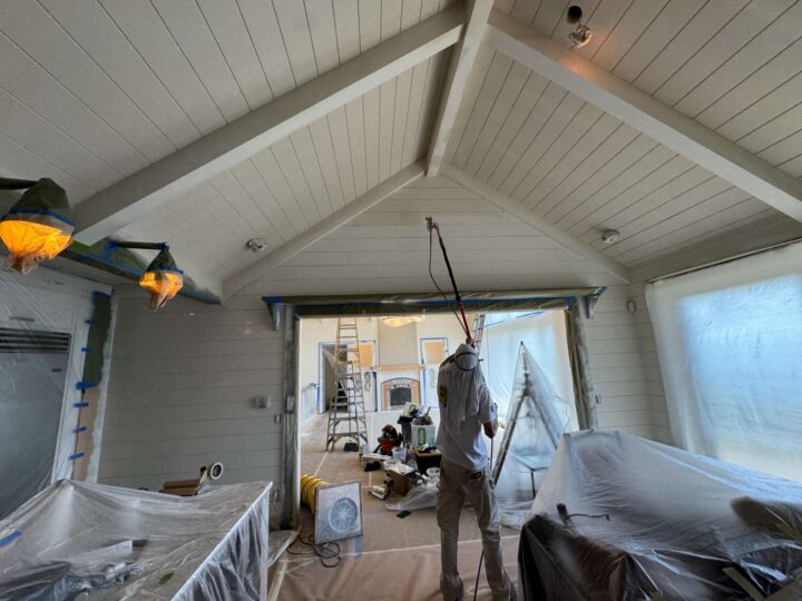 In May 2025, a painter in Portland uses a spray gun to paint an interior ceiling, covering furniture and surfaces with plastic sheets as advised by Duffy of Pearl Painters.