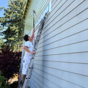 In May 2025, a painter from Pearl Painters uses a roller and ladder to paint the upper exterior wall of a house in Portland light green.