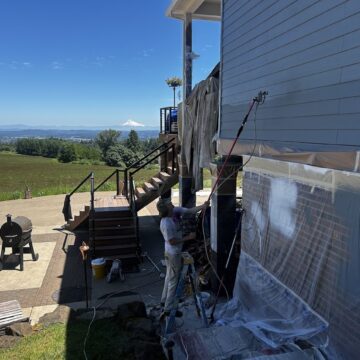 A painter from Pearl Painters uses a sprayer to paint the exterior siding of an Anderson home, with plastic sheets protecting nearby surfaces. The work takes place near Portland, with a scenic landscape and mountain visible in the background on a sunny June 2025 day.