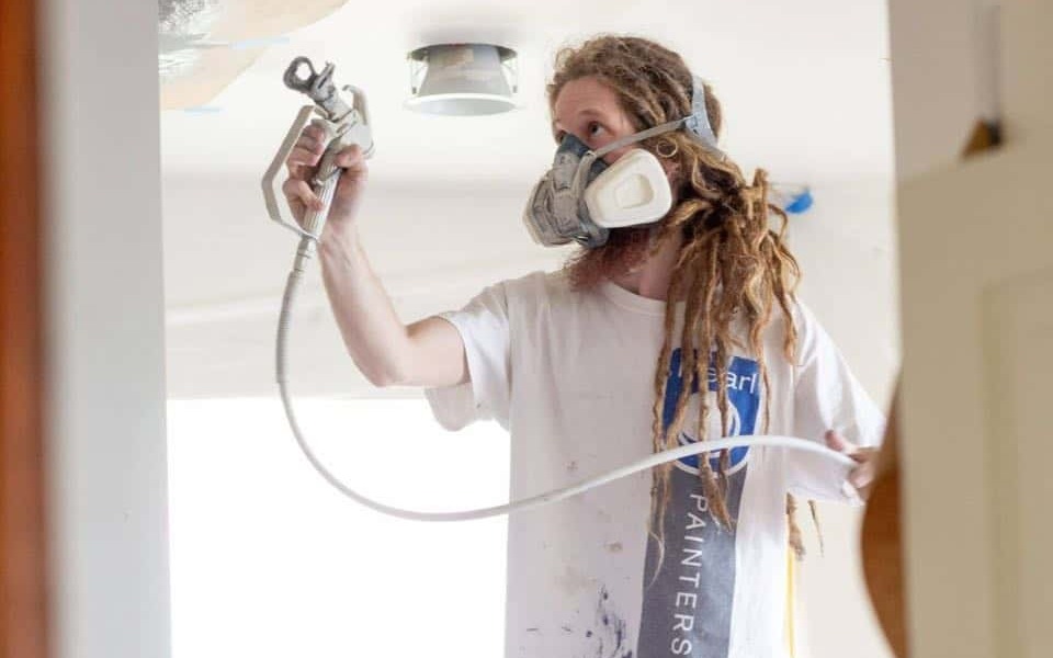 Person wearing a respirator mask and holding a paint sprayer, efficiently spraying the ceiling indoors while ensuring the nearby interior wood trim is protected.