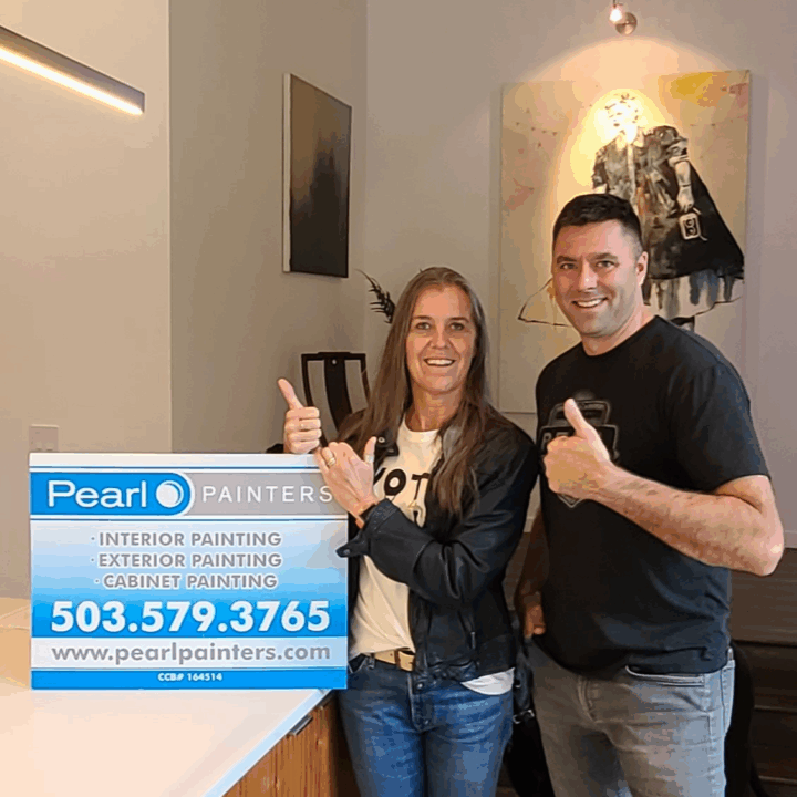 Two people indoors give thumbs up by a Pearl Painters sign showing painting info; artwork and lights in background, Portland.
