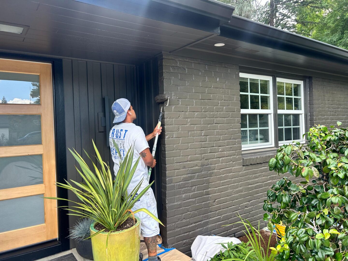 Painter uses a roller to paint the exterior brick wall of a house near the front door and window, with plants nearby. Pearl Painters painting in Portland.