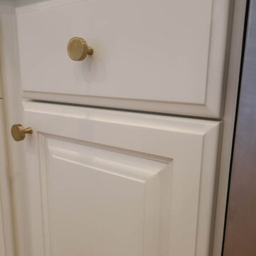 White kitchen cabinet with a drawer and door, featuring gold knobs, set beneath a dark stone countertop.